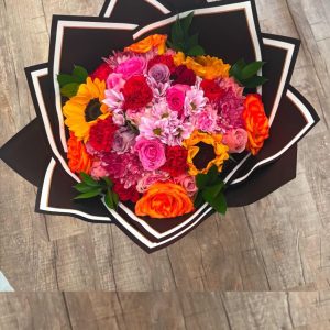 Colorful mixed flower bouquet with sunflowers, roses, assorted blooms, greenery, and black wrapping with light trim.